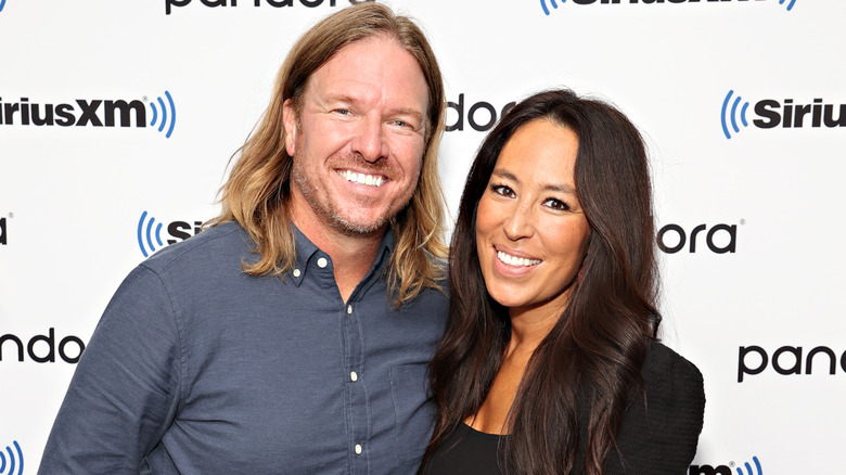 Joanna Gaines smiling in a black top with long loose hair with Chip Gaines in gray button down shirt