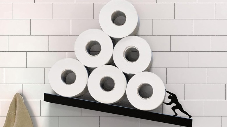 A toilet paper holder that is slanted, showing a black figure pushing the toilet rolls to keep them from falling off