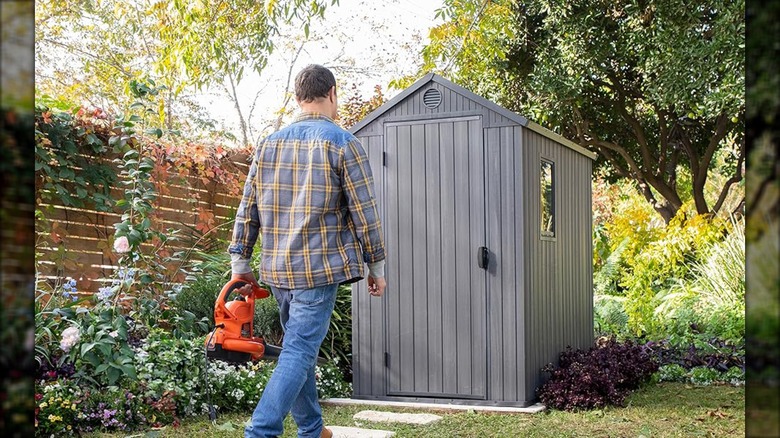 A man carries a leaf blower toward a modern grey shed