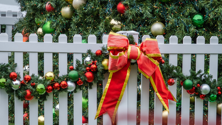 Christmas ornaments hanging on a white picket fence with a Christmas tree behind it