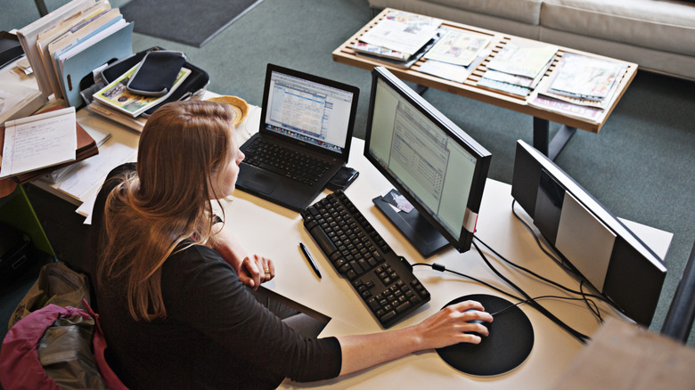 A woman sitting at a desk with three monitors and several documents and files