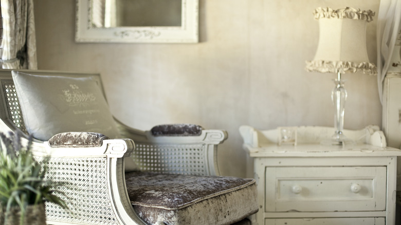 French cane chair with velvet upholstery next to shabby-chic dresser with lamp and ruffle shade