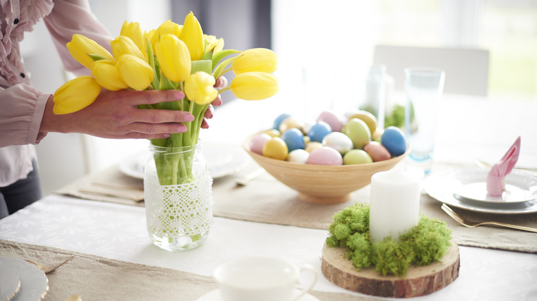 A woman placing yellow tulips in a vase on a table decorated for Easter.