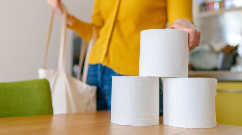 Woman with shopping tote bag on arm stacking three toilet paper rolls on dining table