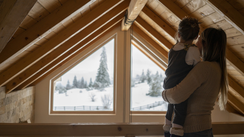 a mother and child standing in an attic and looking out the window at snow covered trees and landsacpe