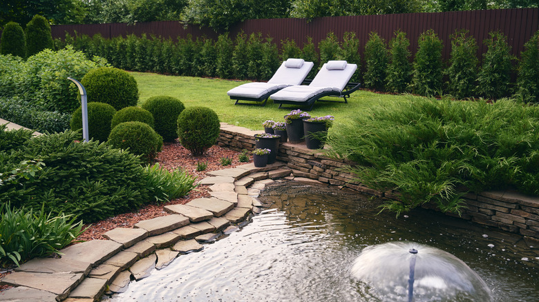 A serene and relaxing garden space with two reclining lounges flanked by hedging and a pond with a fountain in the foreground