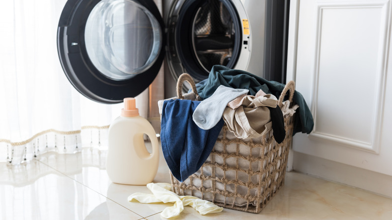 Open laundry machine with a basket of clothes and bottle of detergent sitting in front