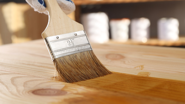 A gloved hand paints a light stain onto a piece of wood.