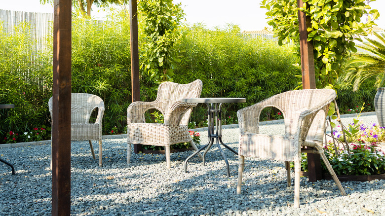 A gravel patio is decorated with wooden pergola, a table, and white chairs