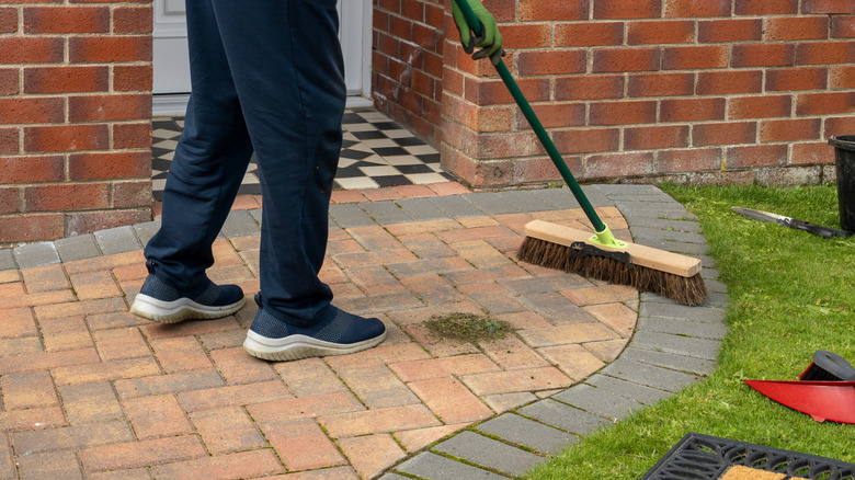 A person in sweatpants and sneakers uses a broom to clean their brick walkway.