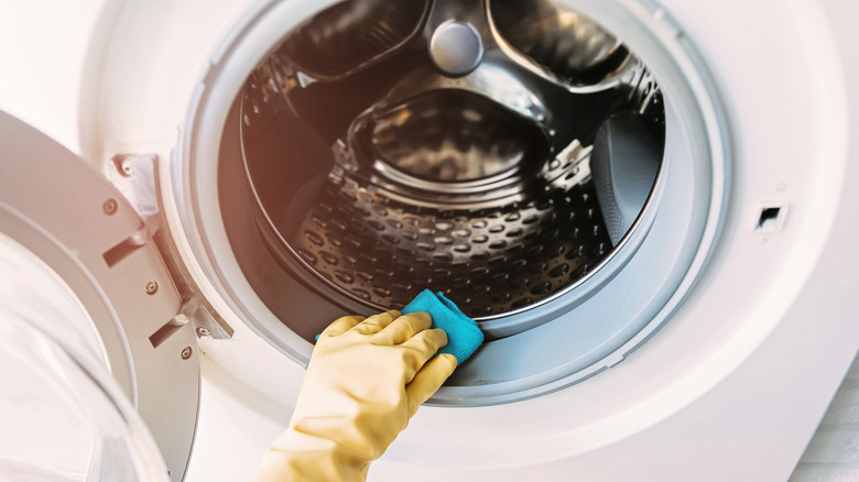 A hand in a yellow cleaning glove is wiping a dirty washing machine drum down with a blue cloth