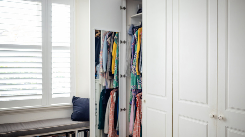 Small closet with clothes and white cabinets with sunlight streaming in from a window