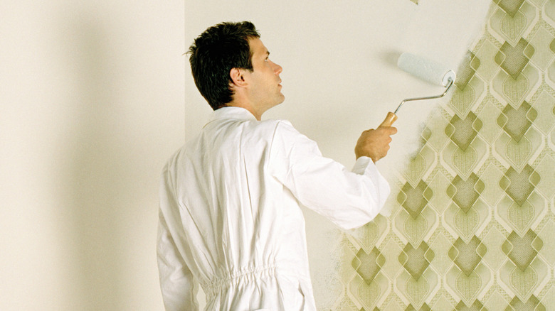 man in white jumpsuit paints white over green patterned wallpaper