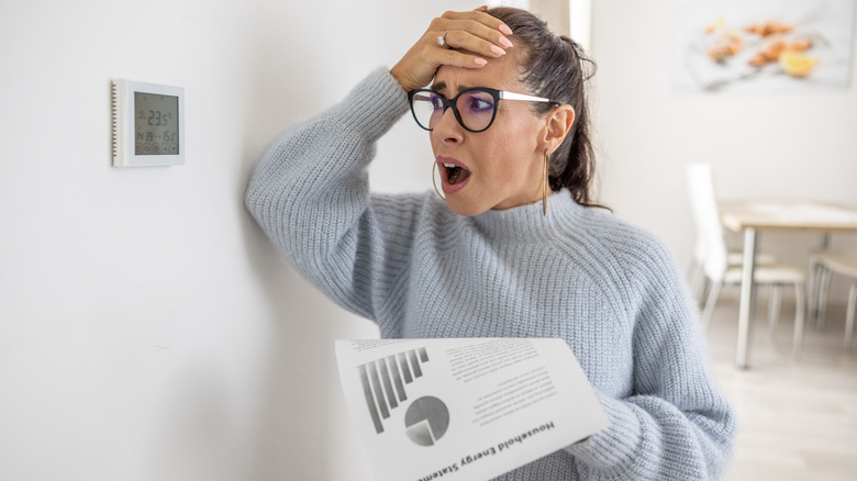 Woman looks at thermostat with mouth open and hand on head