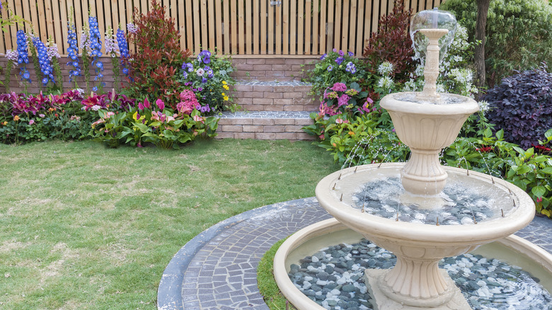 A multi-layered fountain sits in the corner of a yard that features colorful flowers and bushes.