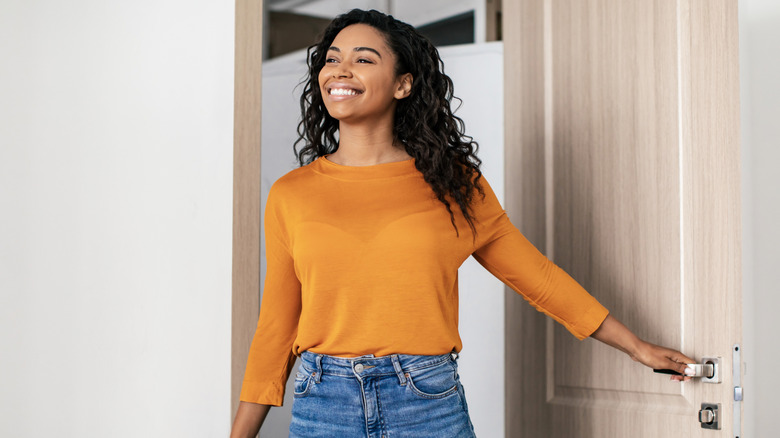 Woman smiling and walking through front door with white empty wall beside her