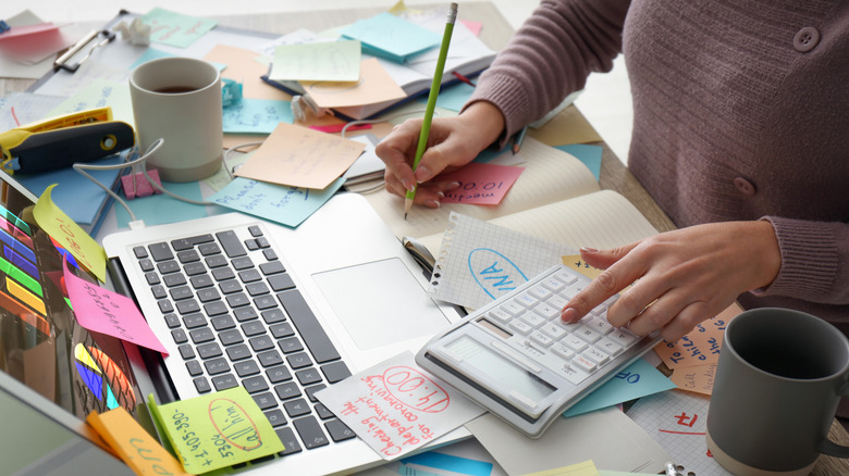 A woman writes in a notebook on a messy and cluttered desk.