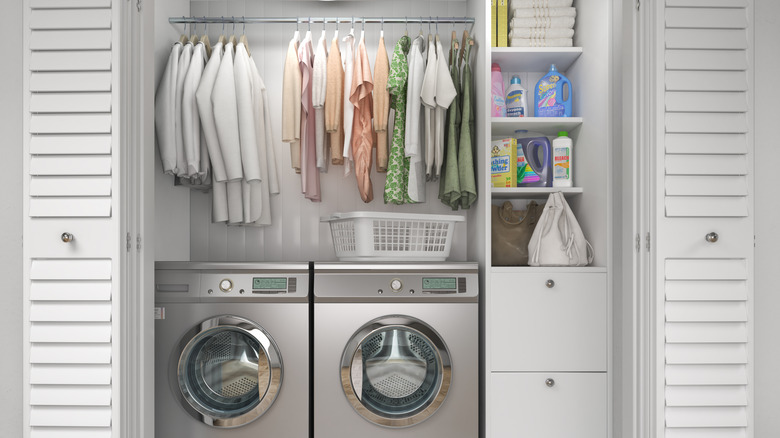 Closet with washing machines, laundry accessories and hanging clothes.