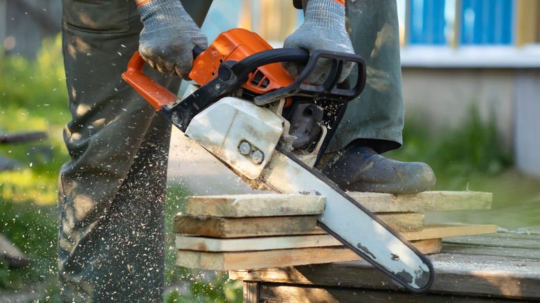 A man using a chainsaw to cut wood planks, he is stepping on the planks using his left leg