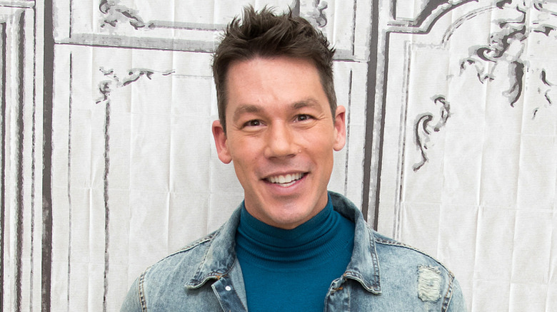 David Bromstad in blue turtle neck and jean jacket smiling in front of wall