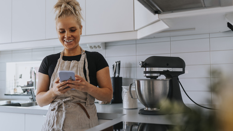 Woman standing in a kitchen using her cellphone with a few kitchen appliances on the counter