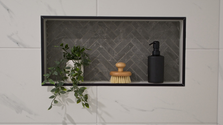 A dark chevron tiled shower niche in a white marble tiled shower with a plant, brush, and dispenser on the shelf.