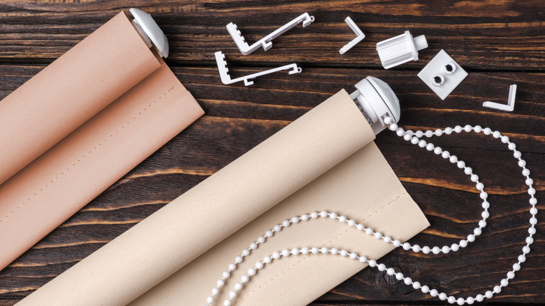 Two roller shades with beige fabric and hanging hardware placed on a wood surface