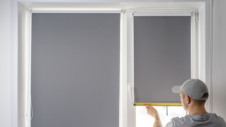 A man in a gray hat measuring the width of the window below a blackout shade