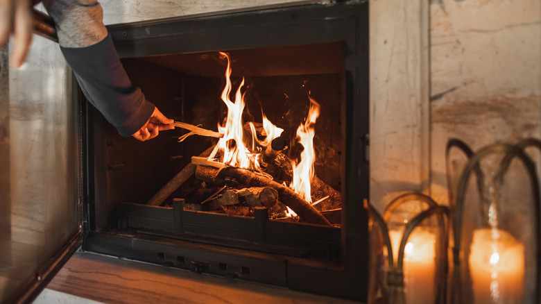 A person carefully places a piece of wood into a lit fireplace