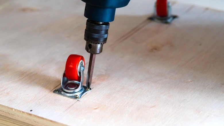 drill screwing small red caster wheel into plywood base