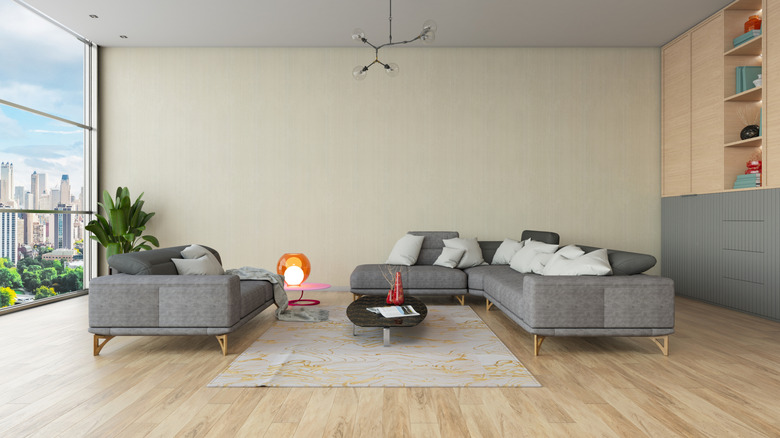 Living room set up with modern couches in front of an empty wall, floor-to-ceiling glass windows on one side and shelving unit on the other.