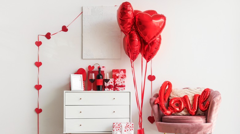 A chair and chest of drawers are covered in red Valentine's Day decorations, including heart balloons and gifts.