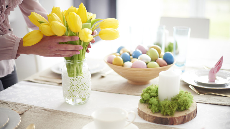 Person placing yellow tulips in glass jar on table decorated for Easter