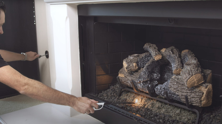 A man turns a fireplace valve key while using a long lighter to ignite a fire