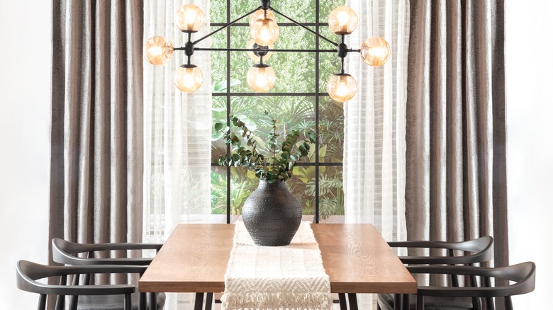 A pendant light fixture hangs over a modern table with a large dark vase in the middle atop a white runner.