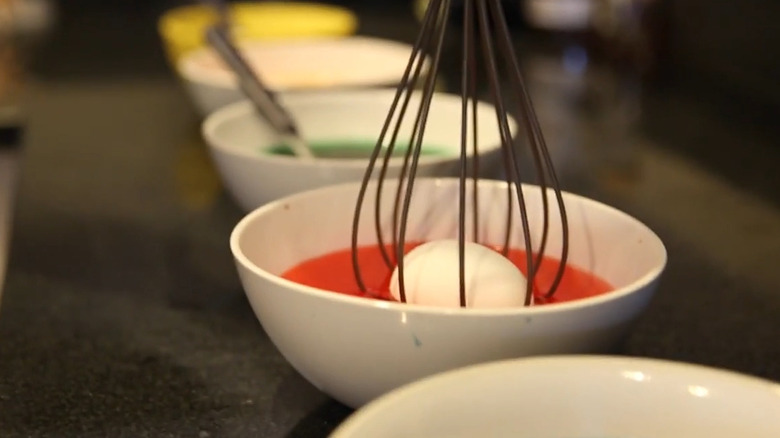 Balloon whisk dipped into bowl of red food dye with egg