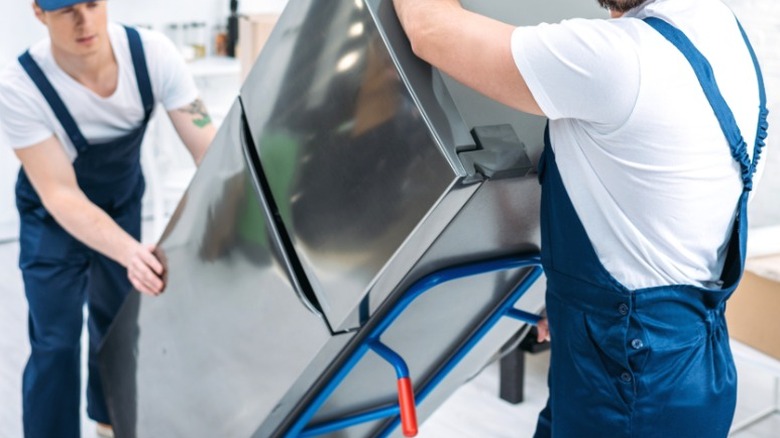 movers in blue caps and overalls transporting small top-freezer refrigerator on handtruck