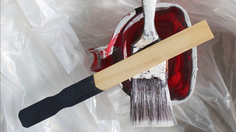 paint stir stick with black paint stain sitting on used brush and paint pail on plastic drop cloth