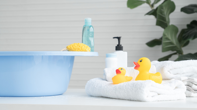 Blue baby bathtub with yellow sponge, blue soap bottle, in room with green plant and towels decorated with yellow rubber duckies