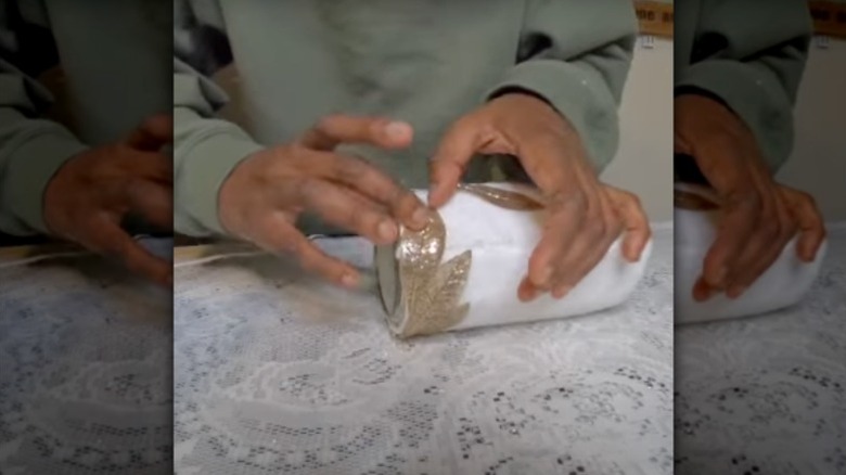 A woman holds a vase she made using faux foliage and a white microfiber cloth wrapped around a jar