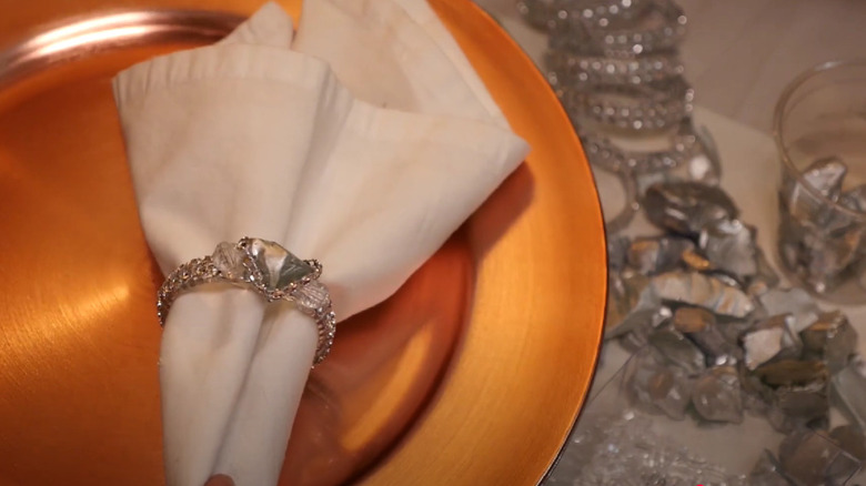 An DIY napkin ring and napkin setting sitting on a gold charger plate with stones and rings in the background.