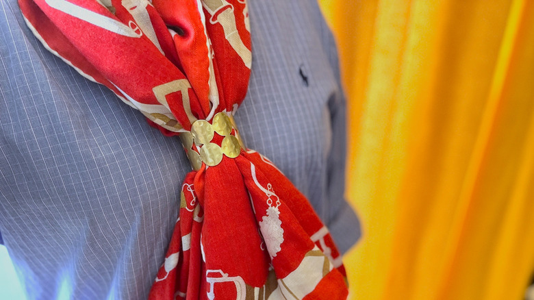 A person standing in front of a yellow background wearing a blue shirt and red scarf held together by a gold ring.