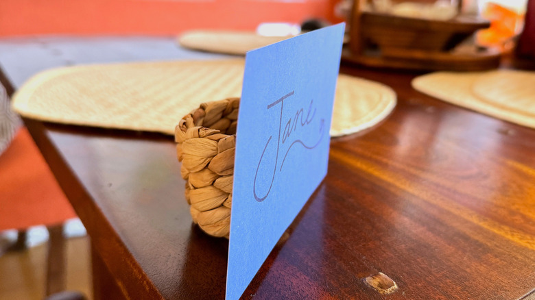 A straw napkin holder attached to a blue name card sitting on a table.