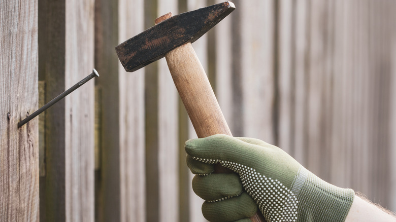 Gloved hand driving a nail into a wooden fence with a hammer