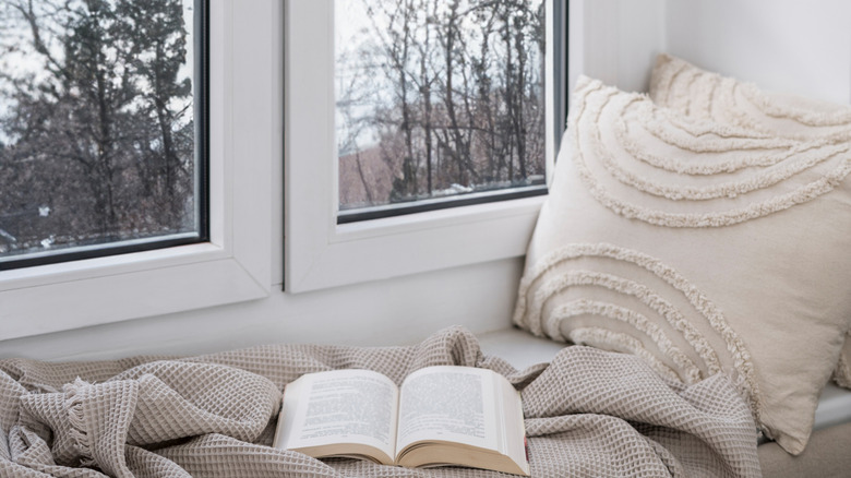 A window seat adorned with a pillow, blanket, and book offers a view of the cold and snowy winter's day outside.