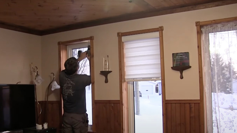 A man installs zebra blinds on the tall, thin windows in his living room