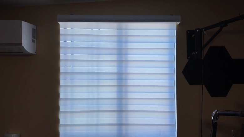 Light beams through gaps between the stripes of a white zebra blind installed on a small window.