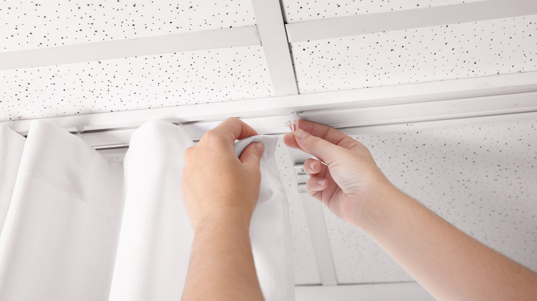 A person hangs sheer white curtain to a ceiling mounted curtain track