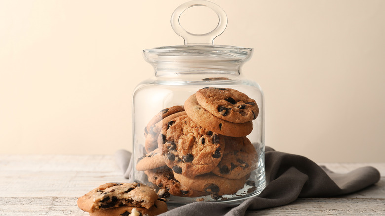 Clear cookie jar filled with chocolate chip cookies sits on a wood surface