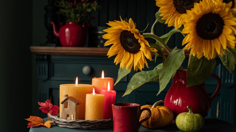 A vase of sunflowers is on a table with candles and gourds.